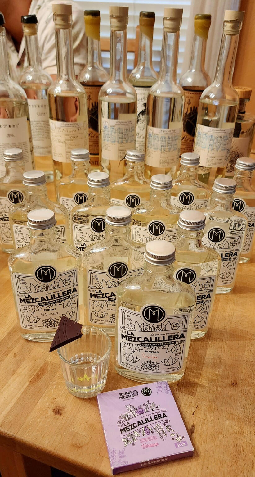 Something new to explore A collection of La Mezcalillera and other agave spirit bottles on a wooden table, with a tasting glass and chocolate in the foreground.